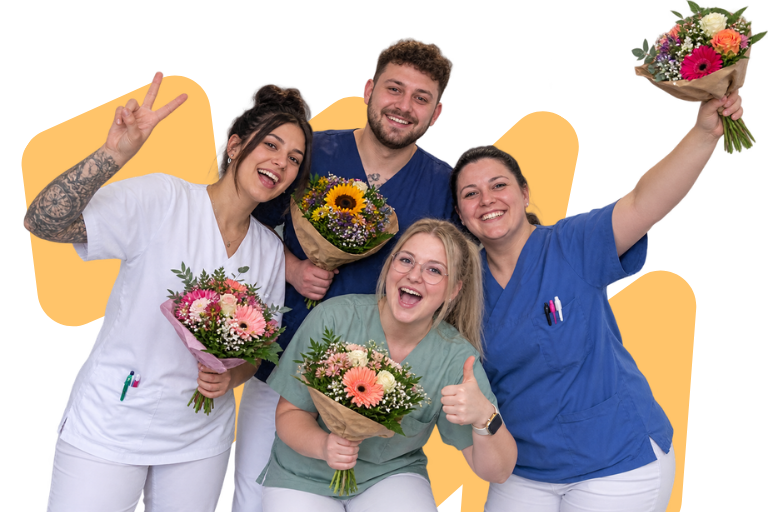 Vier Pflegekräfte in Kasacks halten Blumensträuße in die Höhe und freuen sich über ihre bestandene Weiterbildung
